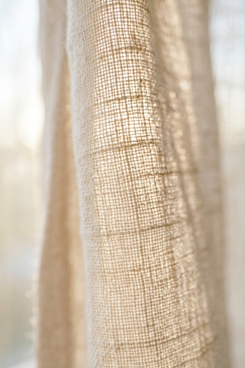 Close-up of natural linen curtain fabric backlit by soft morning daylight, showing the texture of the weave filtering warm light through the fibres.