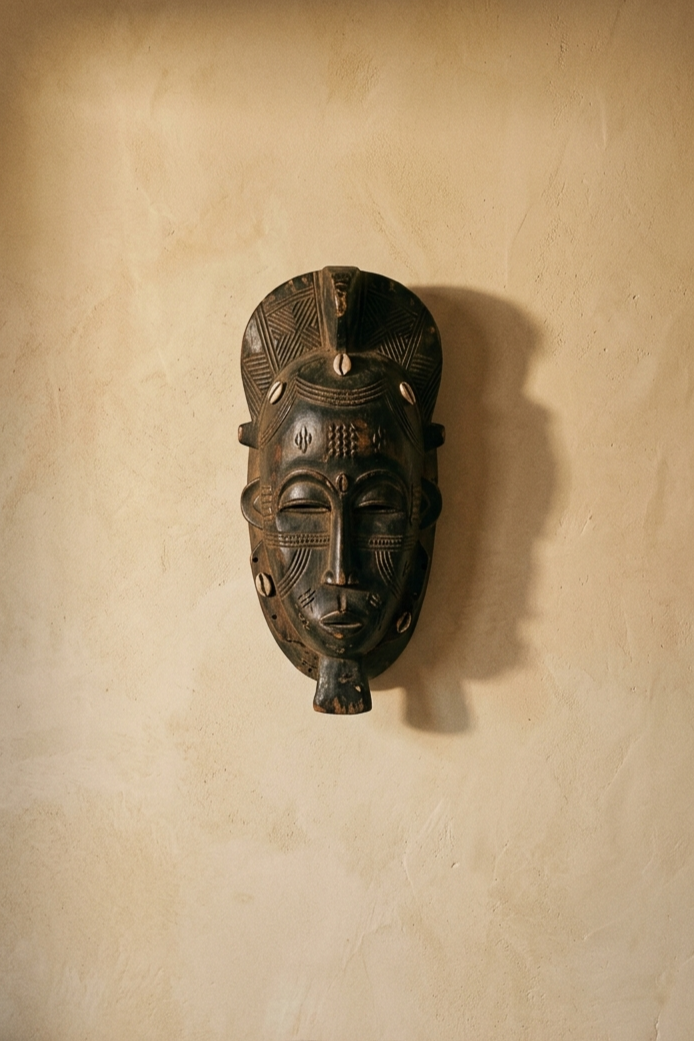 A single hand-carved wooden sculptural object mounted alone on a warm parchment wall with generous space around it, lit by directional warm light with the edge of a linen sofa just visible below